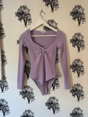 Ljuslila ribbad body med rosett - Trendig ljuslila body med lång ärm och ribbad struktur. Djup v-ringning framtill och en söt rosettdetalj vid bysten. Perfekt att styla till både jeans och kjol för en snygg look.