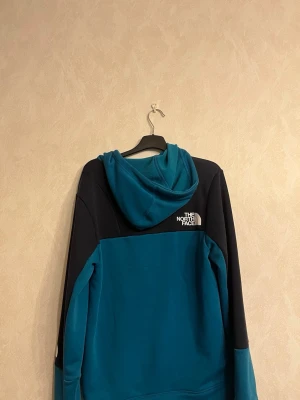 Blå och marin hoodie från The North Face - Snygg hoodie från The North Face i tvåfärgad design med marinblå upptill och ärmar samt klarblå nedtill. Stor luva med dragsko och stor magficka framtill. Logga på bröstet och ärmen. Perfekt för en avslappnad streetwear-look.