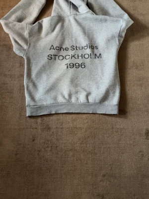 Grå hoodie Acne Studios Stockholm - Snygg grå hoodie från Acne Studios med tryck 'Acne Studios STOCKHOLM 1996' på ryggen. Klassisk passform med huva och ribbade muddar vid ärmslut och nederkant. Perfekt för en avslappnad och trendig look.