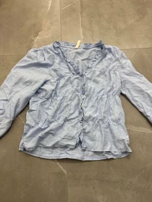 Ljusblå volangblus med knappar - Söt ljusblå blus med volangdetaljer och knäppning framtill. Blusen har v-ringning och långa ärmar, samt en lätt rynkad passform som ger en avslappnad look. Perfekt att matcha med jeans eller kjol för en trendig stil.