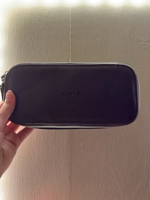 Brun mini necessär från CAIA i läderimitation - Säljer en stilren brun plånbok från CAIA i läderimitation. Plånboken har en avlång, rektangulär form och stängs med dragkedja runtom. Diskret CAIA-logga på framsidan. Perfekt för att hålla ordning på kort och kontanter.