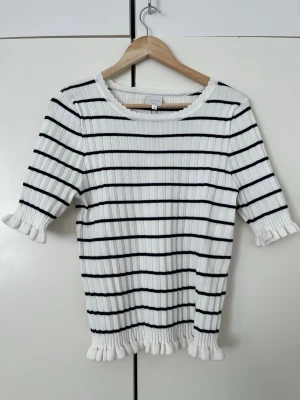 Vit ribbad topp med svarta ränder - Snygg vit ribbad topp från Mingel med svarta horisontella ränder. Toppen har volangdetaljer vid ärmslut och nederkant samt rund halsringning. Materialet är stretchigt och ger en bekväm passform. Perfekt för en solig dag! Varan har ej används då prislappen är kvar. Men den har en fläck på den vänstra ärmen baktill när jag köpte toppen. Inget man ser framefrån! Den kostade 599kr och jag säljer den för 249kr. Du kan alltid höra av dig så kan vi diskutera priset!