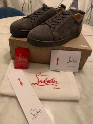 Gråa Christian Louboutin sneakers - Snygga gråa sneakers från Christian Louboutin i mocka med klassisk röd sula. Lågt skaft, rund tå och matchande grå skosnören. Kommer med originalkartong, dustbag och certifikat. Perfekta för dig som vill ha något exklusivt och stilrent.