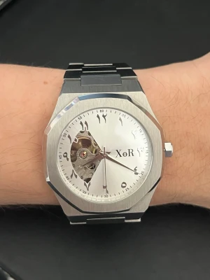 XoR Obsucra Automatic 41mm Silver - Stilren klocka från XoR med silverfärgad urtavla och armband i stainless steel. Urtavlan har arabiska siffror och ett öppet fönster som visar mekaniken inuti. Klockan är använd men utseende vis ser den fortfarande ny ut. Kommer i originalbox. 