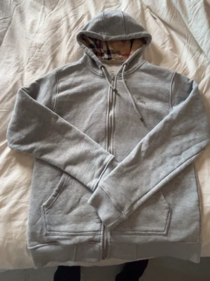 Grå zip hoodie från Burberry - Snygg grå zip hoodie från Burberry med klassiskt rutigt foder i huvan och broderad logga på bröstet. Hoodien har dragsko i huvan, två fickor fram och ribbade muddar. Perfekt för en avslappnad och stilren look.