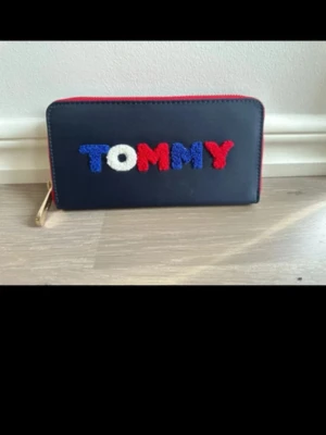 Mörkblå Tommy Hilfiger plånbok - Snygg mörkblå plånbok från Tommy Hilfiger med färgglada bokstäver i rött, vitt och blått på framsidan. Plånboken har dragkedja i guld och röd kant. Tillverkad i slätt konstläder och har en stilren, rektangulär form.