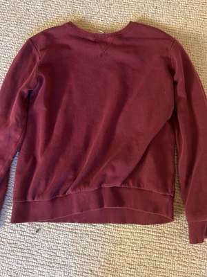 Vinröd tjocktröja  - En stilren bordeaux crewneck sweatshirt med rund hals och ribbade muddar vid ärmslut och midja. Perfekt för en avslappnad look. Materialet ser ut att vara bomull eller bomullsblandning. Enkel och klassisk design utan tryck eller logga.