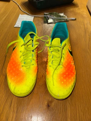 Nike neon-gula fotbollsskor - Säljer ett par riktigt snygga fotbollsskor från Nike i neon-gult med orange detaljer och svart swoosh på sidan. Skorna har en texturerad ovandel med små dobbar för extra grepp och kontroll. Insidan är turkosblå för en cool kontrast. Perfekta för dig som vill sticka ut på planen!