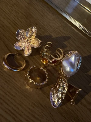 Sju trendiga guldringar med detaljer - Säljer ett set med sju unika guldfärgade ringar i olika former. En ring har en stor blomma, en annan har en stor pärlliknande detalj, och flera har vågiga, strukturerade eller stenar i olika färger. Perfekta för att mixa och matcha för en personlig stil.