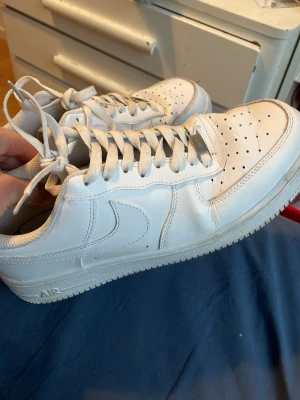 Vita Nike Air Force 1 sneakers - Klassiska vita Nike Air Force 1 sneakers i lågt utförande. Skorna har en stilren design med perforerad tå, vita skosnören och den ikoniska swoosh-loggan på sidan. Tillverkade i läder med gummisula för bra grepp. Perfekta för en clean och tidlös look.