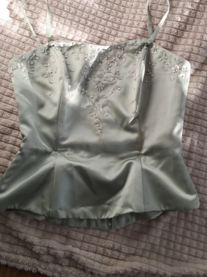 Ljusgrön korsettopp med broderi och kjol - Supersnygg ljusgrön korsettopp i satin med tunna axelband och vackra broderade blommor framtill. Toppen har figurnära passform och ger en elegant siluett. Perfekt för dig som vill sticka ut med en unik och feminin look. Kjol ingår