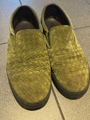 Bottega Ventea Olivgröna slip-on sneakers i mocka - Säljer ett par olivgröna slip-on sneakers med flätad ovandel i mocka. Skorna har svart sula och diskret broderad detalj bak. Perfekta för dig som gillar stilrena och unika sneakers med skön passform. Passar 42/43