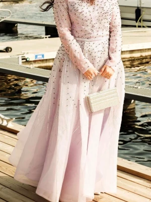 Ljuslila långklänning med pärlor - Prinsess ljuslila långklänning med långa ärmar. Klänningen är dekorerad med glittriga pärlor och stenar i ett stjärnformat mönster över hela plagget. Använt en gång på balen.