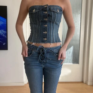 Jeans korsett topp med knappar - Säljer en snygg jeans korsett topp i blå denim med knappar framtill och råa kanter. Toppen är axelbandslös och har en figurnära passform som framhäver midjan. Perfekt för att skapa en cool och trendig look. Använd en gång, från Zara. 