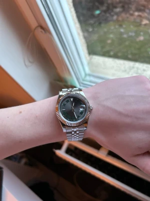 Silverfärgad klocka med grön urtavla - Stilren klocka i rostfritt stål med silverfärgad länk och grön urtavla. Klockan har romerska siffror, räfflad bezel och datumfönster. Klassisk design som passar till många stilar.