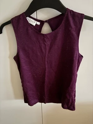 Glittrig mörklila ärmlös topp - Snygg mörklila ärmlös topp med glittriga detaljer i tyget. Toppen har rund halsringning och en liten knapp i nacken för stängning. Perfekt för dig som vill sticka ut lite extra.