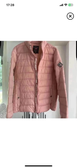 Rosa pufferjacka från Polo Club - Säljer en ljusrosa pufferjacka från Polo Club med hög krage och quiltad design. Jackan har dragkedja framtill, två fickor med tryckknappar och en patch på ärmen. Materialet är lätt och vadderat, perfekt för kalla dagar.