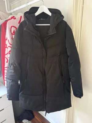 Svart lång pufferjacka med huva från ZARA - Säljer en svart lång pufferjacka från ZARA med stor huva och två dragkedjeförsedda fickor framtill. Jackan har en rak passform och grova sömmar som ger en klassisk pufferlook. Perfekt för kalla vinterdagar och riktigt mysig att bära.