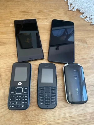 Blandat mobilpaket med 5 mobiler - Säljer ett paket med fem olika mobiltelefoner: en svart iPhone, en svart Sony Xperia, två klassiska knappmobiler från Nokia och On samt en svart och vit Doro viktelefon. Perfekt för dig som gillar äldre mobiler eller behöver reservdelar. Alla är i svart eller svart/vit färg.
