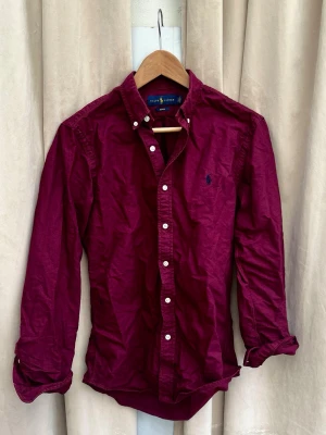 Vinröd skjorta från Ralph Lauren - Stilren vinröd skjorta från Ralph Lauren. Skjortan har vita knappar och den ikoniska broderade loggan på bröstet. Tillverkad i mjuk bomull som känns skön mot huden. Perfekt för när man ska på middag eller att ha till vardags men fortfarande lite uppklädd. Hör av er vid frågor!👊