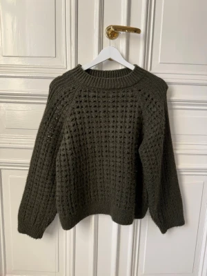 Olivgrön stickad tröja med hålmönster - Säljer en olivgrön stickad tröja med hålmönster och bred ribbad kant vid hals, ärmslut och nederkant. Tröjan har en avslappnad passform och långa ärmar. Perfekt för lager på lager och riktigt mysig till hösten.
