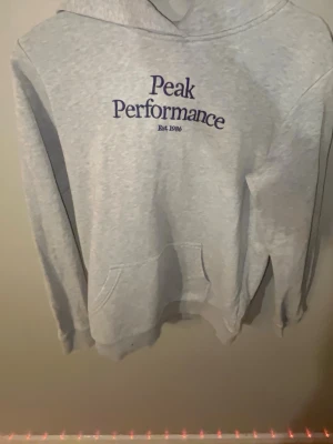 Grå hoodie från Peak Performance - Snygg ljusgrå hoodie från Peak Performance med mörkblå text på bröstet och känguruficka framtill. Klassisk huva och mjukt material som känns skönt mot huden. Perfekt för chill dagar eller när du vill ha en clean look.