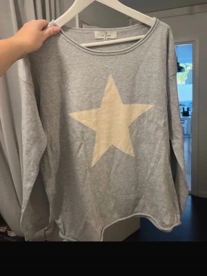 Grå tröja med stjärna - Grå långärmad tröja  med en stor vit stjärna på bröstet. Tröjan har en avslappnad passform och rund halsringning. Perfekt för en chill och enkel stil. Kolla HÅLET vid armen men det går nog att laga!