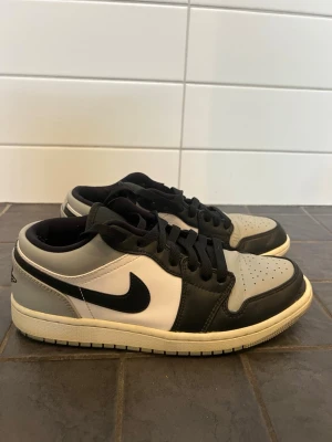 Air Jordan 1 Low svart/vit/grå - Säljer ett par Nike Air Jordan 1 Low sneakers i svart, vitt och grått läder. Klassisk siluett med svart nikemärke, vita paneler och grå detaljer. Lågt skaft och vita sulor.  Saknar plast längst fram på ett av skosnörerna, se bild. 
