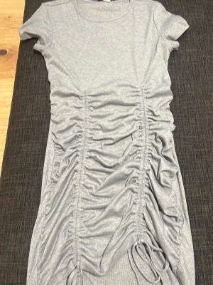 Grå ribbad klänning med rynkdetaljer - Trendig grå klänning i ribbat material med korta ärmar och rund halsringning. Klänningen har snygga rynkade detaljer framtill och justerbara snören längst ner för en cool look. Perfekt för dig som gillar en modern och avslappnad stil.