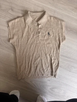 Beige kabelstickad polotopp från Ralph Lauren - Kabelstickad beige polotopp från Ralph Lauren med korta ärmar och klassisk krage. Toppen har en liten broderad logga på bröstet och är tillverkad i mjukt material som känns lyxigt. Perfekt för en clean och stilren look.