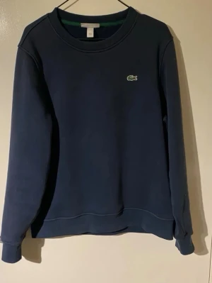 Mörkblå sweatshirt från Lacoste - Klassisk mörkblå sweatshirt från Lacoste med rund halsringning och den ikoniska gröna krokodilloggan broderad på bröstet. Tröjan har ribbade muddar vid ärmslut och nederkant. Perfekt för en avslappnad och stilren look.