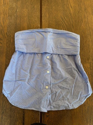 Blåvit randig tubtopp med knappar S Zara  - Snygg blåvit tubtopp med tunna vertikala ränder och knappar framtill. Toppen har en bred kant upptill och är gjord i ett lätt bomullstyg. Perfekt för en avslappnad och trendig look.