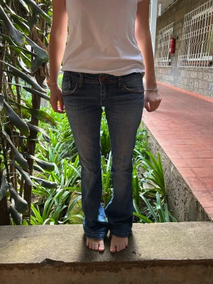 Blå bootcut jeans med låg midja - Snygga blå bootcut jeans med låg midja och klassiska fem fickor. Jeansen har en lätt tvättad look och subtila slitningar för en avslappnad stil. Passar perfekt till sneakers eller klackar och ger en tidlös vibe till din outfit.