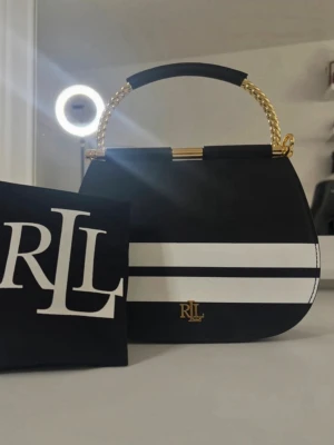 Svart och vit handväska från Ralph Lauren - Snygg svart handväska i läder från Lauren Ralph Lauren med två vita ränder på framsidan och guldfärgad logga. Väskan har ett elegant, rundat handtag i guldfärgad metall och svart läderdetalj. Perfekt accessoar för att lyfta din outfit.
