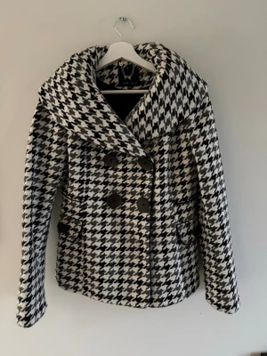 Höstig kappa - Snygg svartvit kappa från Amisu i klassiskt hundtandsmönster. Dubbelknäppt framtill med stora svarta knappar och bred krage. Storlek passar xs-m och är i bra skick.