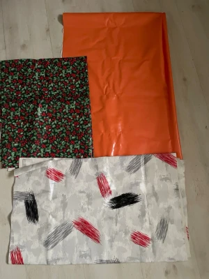 Tre olika Kraftiks Vaxdukar i blandade mönster - Säljer tre kraftiga Vaxdukar : ett orange enfärgat, ett med Lingonmönster i rött och grönt på svart botten, samt ett vitt tyg med abstrakta penseldrag i svart, rött och grått. Perfekt för DIY-projekt, sömnad eller pyssel.