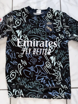 Real Madrid tränings t-shirt - Snygg svart Real Madrid tränings t-shirt från Adidas med grafitti-inspirerat mönster i blått och vitt. Stort Real Madrid-märke på bröstet och 'Emirates Fly Better' tryckt framtill. Tillverkad i lätt och ventilerande material, perfekt för träning eller match. Den är i storlek 152 och passar för barn. 