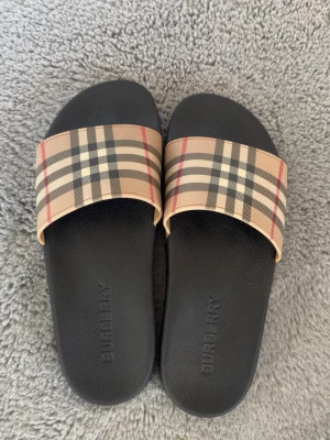 Burberry sandaler med rutigt band - Snygga sandaler från Burberry med klassiskt beige, svart och rött rutigt band över foten. Sulan är svart och har Burberry-loggan präglad. Perfekta för sommaren och ger en lyxig touch till din outfit.