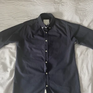 Mörkblå skjorta från 157, storlek M - Snygg mörkblå skjorta från 157 i klassisk modell med knappar hela vägen och button down-krage. Skjortan är långärmad och tillverkad i mjuk bomull, perfekt för en clean och stilren look.