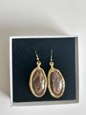 Ovala örhängen med brun sten - Använt två gånger. De är snygga ovala örhängen med en stor brun sten i mitten, omgivna av en guldfärgad metallram med hamrad struktur. Krokar i metall för enkel användning. Perfekta för dig som gillar unika accessoarer med vintagekänsla.