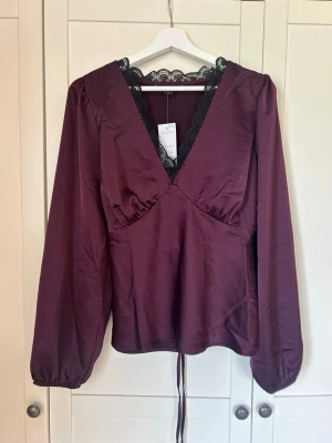 Vinröd blus med spetsdetalj - Vinröd blus med djup V-ringning och svarta spetsdetaljer vid halsen. Långa puffärmar med resår vid ärmslut och knytband i midjan. Aldrig använt då jag köpte fel storlek, köpt från Zalando, märket är New Look. Är ganska lite liten i storleken så passar nog 42-46! 