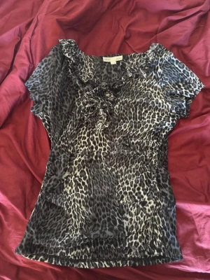 Leopardmönstrad blus från Cleo - Snygg leopardmönstrad blus från Cleo i svart, grått och vitt. Blusen har korta ärmar, volangdetaljer vid halsringningen och en dekorativ knytning framtill. Tillverkad i ett lätt och luftigt material som känns skönt mot huden.