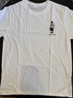 Vit t-shirt med tryck och text NICE - Vit T-shirt med NICE-tryck – streetwear (M/L)  Vit T-shirt i mjuk bomull med ett iögonfallande tryck på ryggen med texten “NICE” och en tecknad figur. Framtill finns en mindre illustration med texten “NOW I GLEANED”. Klassisk rund hals och korta ärmar ger en bekväm passform. Perfekt för en avslappnad och trendig streetwear-look som är lätt att matcha med olika outfits.  Storlek: M–L  💬 Hör av dig vid intresse!