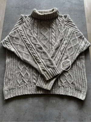 Grå kabelstickad polotröja - L - Säljer en grå kabelstickad polotröja med höga muddar och klassiskt flätmönster. Tröjan har en bred polokrage och är gjord i ett mjukt garn som känns skönt mot huden. Perfekt för kyliga dagar!
