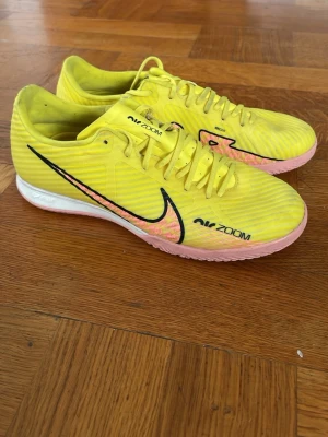 Nike Air Zoom gula fotbollsskor - Snygga fotbollsskor från Nike Air Zoom i en fräsch gul färg med svarta detaljer och rosa sula. Skorna har snörning och ovandel i syntetmaterial för bra känsla på planen. Perfekta för dig som vill sticka ut på futsal eller inomhusplanen.