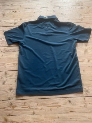 Mörkblå FootJoy pikétröja - Mörkblå pikétröja från FootJoy med klassisk krage och två knappar framtill. Tröjan har korta ärmar och är tillverkad i ett lätt, sportigt material. FootJoy-logga syns på kragen både fram och bak för en clean och sportig look.