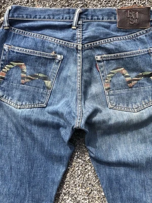 Blå jeans från Evisu med camodetalj - Säljer ett par blå jeans från Evisu med unika camoinspirerade detaljer på bakfickorna och klassisk läderpatch i midjan. Jeansen har rak passform och tydliga sömmar. Perfekta för dig som vill sticka ut med streetwear-vibe.