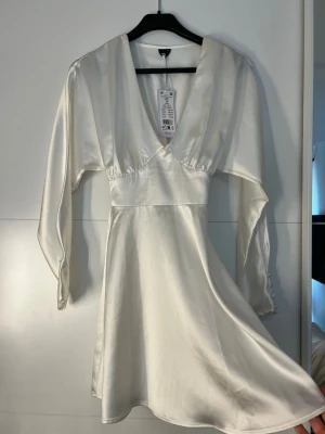 Vit satin klänning med v-ringning - Elegant vit klänning i satin med djup v-ringning och långa ärmar. Klänningen har markerad midja och knappar vid ärmsluten för en extra detalj. Perfekt för dig som vill ha en stilren och chic look.