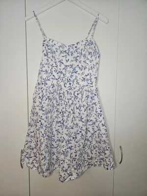 Vit och blå blommig klänning H&M - Supersöt vit klänning från H&M med blått blommönster och smala axelband. Kort modell med lätt utställd kjol och elastisk rygg för skön passform. Perfekt för varma dagar och enkel att styla med sneakers eller sandaler.