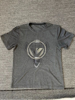 Mörkgrå t-shirt från Armani - Snygg mörkgrå t-shirt från Armani med ett stort silverfärgat logotryck framtill och mindre logga på ryggen. Klassisk rund halsringning och korta ärmar. Tillverkad i mjuk bomull för skön känsla och stilren look.